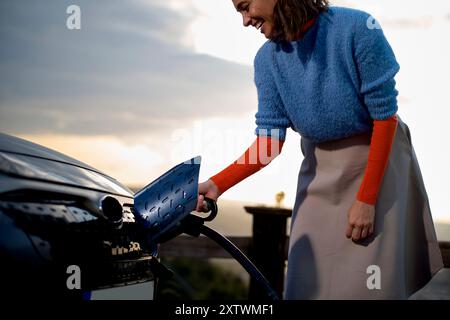 Donna sorridente con maglione blu e gonna che collega un caricabatterie per auto elettriche durante il crepuscolo. Foto Stock