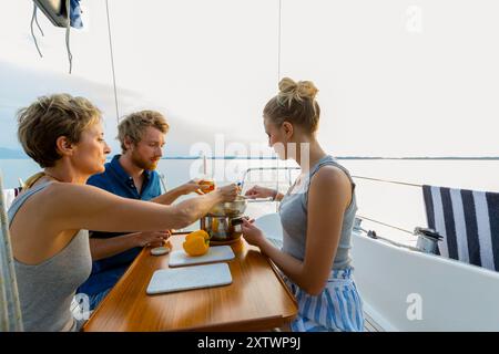 Tre persone si godono un pasto insieme sul ponte di una barca a vela con un tranquillo sfondo d'acqua durante il tramonto. Foto Stock