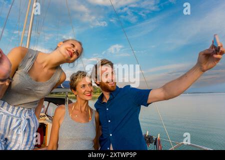 Tre amici sorridenti e scattano un selfie su una barca con un cielo limpido sullo sfondo. Foto Stock