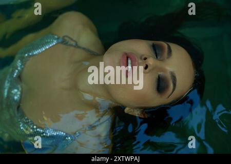 Una donna con i capelli scuri e bagnati galleggia tranquillamente in un corpo d'acqua, indossando un abito scintillante color argento. Foto Stock