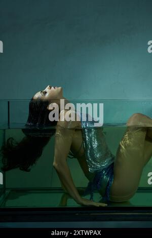 Una giovane donna con capelli mora bagnati galleggia in un acquario, il suo corpo è immerso nell'acqua, indossando un top e una gonna scintillanti. Foto Stock