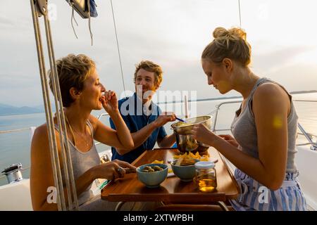 Gli amici si godranno un pasto su una barca a vela al tramonto con un paesaggio acquatico panoramico sullo sfondo. Foto Stock