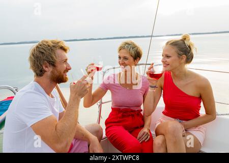 Tre amici si godono un drink e chiacchierano sul ponte di una barca a vela con un tranquillo sfondo d'acqua al tramonto. Foto Stock