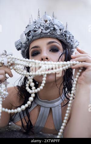 Una giovane donna con i capelli scuri, che indossa una corona di conchiglie e una collana di perle, posa in un corpo d'acqua poco profondo. Foto Stock