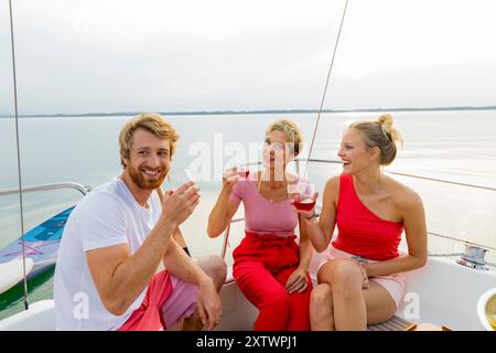 Tre amici si godono un drink su uno yacht con un lago tranquillo sullo sfondo al tramonto. Foto Stock
