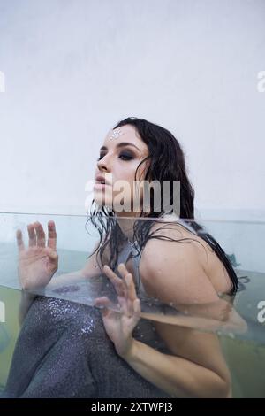 Una giovane donna con lunghi capelli scuri galleggia in un acquario, i suoi occhi pieni di tristezza e desiderio. Foto Stock