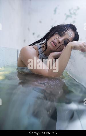 Una giovane donna con lunghi capelli scuri giace in un acquario limpido, il suo volto poggia sul bordo. L'acqua le gira delicatamente intorno. Foto Stock