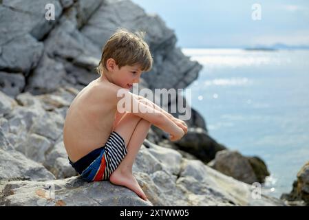 Un ragazzino siede sulla costa rocciosa con il mare sullo sfondo, guardando attentamente in lontananza. Foto Stock