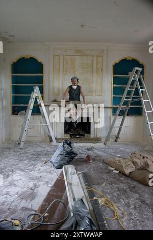 Un giovane uomo siede su un manipolo in una stanza in fase di ristrutturazione con due scale, detriti e strumenti di ristrutturazione intorno. Foto Stock