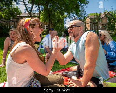 Un uomo e una donna allegri si siedono su una coperta da picnic in un parco e si impegnano in una vivace conversazione circondati da amici in una giornata di sole. Foto Stock