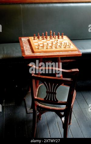 Una scacchiera con i pezzi ordinatamente disposti per un gioco si trova in cima a un tavolo di legno accanto a un divano in pelle nera, con una sedia di legno vuota rivolta verso di esso. Foto Stock