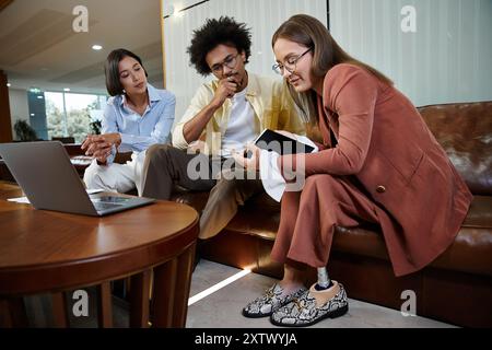 Tre colleghi, tra cui una donna con una gamba protesica, discutono di un progetto su un tablet mentre si siedono su un divano in una moderna hall di ufficio. Foto Stock