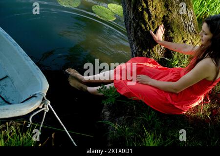 Una donna con un vestito rosso siede accanto a un tranquillo laghetto, immergendosi i piedi nell'acqua, con una barca bianca ormeggiata accanto a lei. Foto Stock