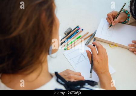 Vista dall'alto, prospettiva di una donna irriconoscibile che disegna su un tavolo con taccuino e matite colorate, ha in mano una matita di grafite, ci sono c Foto Stock