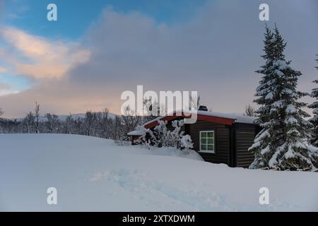 Un piccolo rifugio di caccia innevato sulle montagne norvegesi con tracce di animali in primo piano e vette di montagna sullo sfondo Foto Stock