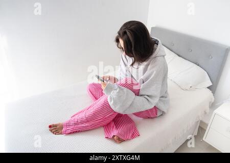 Una giovane ragazza seduta sul letto in una stanza bianca guarda il suo smartphone. Indossa pantaloni rosa e una felpa grigia ed è a piedi nudi. Concetto di d Foto Stock