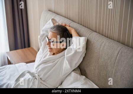 Donna sorridente dai capelli grigi che indossa un accappatoio bianco in camera Foto Stock