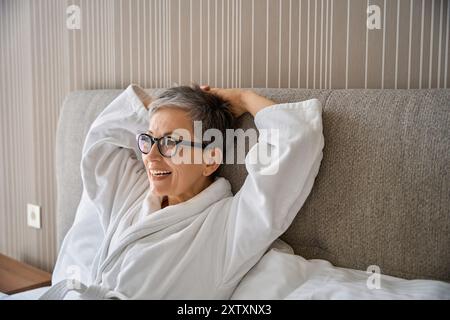 Donna felice dai capelli grigi che indossa un accappatoio bianco in camera Foto Stock