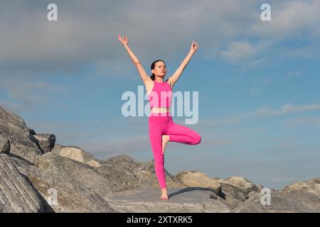 Donna che fa yoga posare un albero in cima a rocce con abbigliamento sportivo rosa Foto Stock