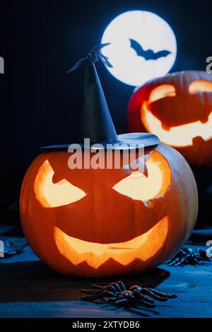 Zucche intagliate luccicanti di Halloween con un sorriso malvagio e un cappello da strega, luna piena e sagoma di pipistrello Foto Stock