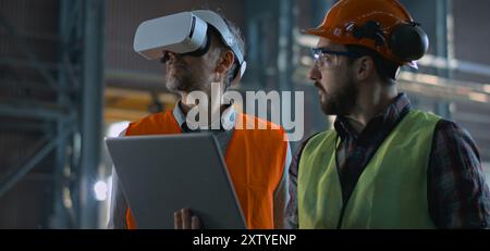 Due tecnici professionisti del settore che indossano l'uniforme di sicurezza e i cappelli rigidi si trovano presso lo stabilimento di produzione e utilizzano laptop e cuffie per la realtà aumentata. Tecnici maturi e dipendenti adulti lavorano in magazzino. Al rallentatore. Foto Stock