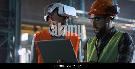 Due tecnici professionisti del settore che indossano l'uniforme di sicurezza e i cappelli rigidi si trovano presso lo stabilimento di produzione e utilizzano laptop e cuffie per la realtà aumentata. Tecnici maturi e dipendenti adulti lavorano in magazzino. Al rallentatore. Foto Stock