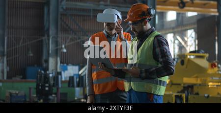 Due tecnici professionisti del settore che indossano uniforme protettiva e caschi si trovano presso lo stabilimento di produzione. Un tecnico maturo utilizza un visore VR, un dipendente adulto lavora su un computer portatile. Al rallentatore. Foto Stock