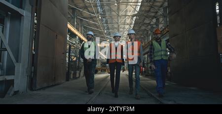 Four heavy industry engineers in safety uniform and hard hats discuss work process walking at manufacturing factory. Mature supervisor holds laptop, talks to diverse workers in warehouse. Slow motion. Foto Stock