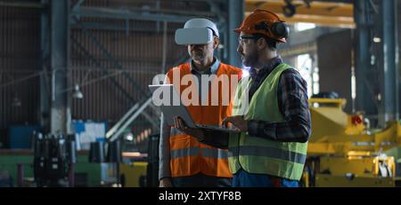 Due tecnici professionisti del settore che indossano uniforme protettiva e caschi si trovano presso lo stabilimento di produzione. Un tecnico maturo utilizza un visore VR, un dipendente adulto lavora su un computer portatile. Al rallentatore. Foto Stock