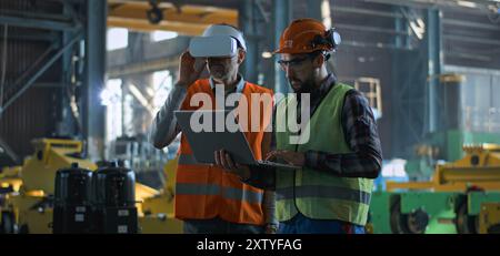 Due tecnici professionisti del settore che indossano uniforme protettiva e caschi si trovano presso lo stabilimento di produzione. Un tecnico maturo utilizza un visore VR, un dipendente adulto lavora su un computer portatile. Al rallentatore. Foto Stock