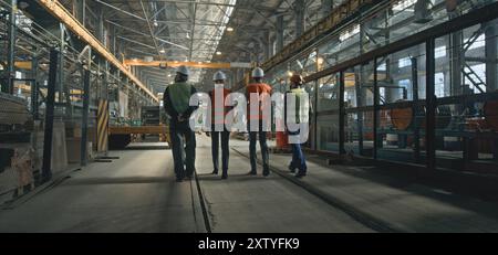 Vista posteriore di quattro tecnici del settore pesante in uniforme e caschi che discutono del processo di produzione, camminando nello stabilimento di produzione. Ispettori maschi e femmine parlano con i dipendenti in fabbrica. Al rallentatore. Foto Stock