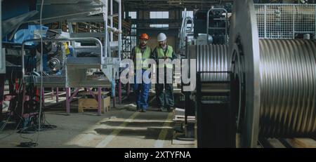 Due tecnici professionisti dell'industria pesante in uniforme protettiva e caschi discutono del processo di produzione che si svolge nello stabilimento di produzione. I dipendenti maschi lavorano nello stabilimento. Al rallentatore. Foto Stock