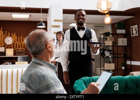 Nella lussuosa hall dell'hotel, il cameriere afro-americano serve caffè agli anziani gentiluomini che visitano la città utilizzando un tablet digitale. Nell'area lounge, il ragazzo anziano in pensione tiene in mano il dispositivo e accetta una tazza di tè dal dipendente. Foto Stock
