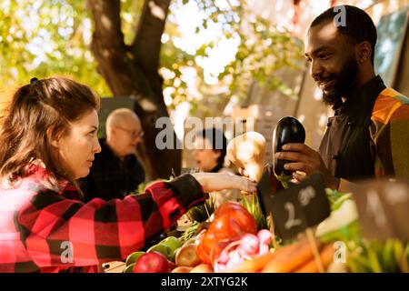In un mercato agricolo locale, un uomo afroamericano si trova dietro un bancone che vende verdure di stagione biologiche fresche a una cliente femminile. Giovane donna che acquista prodotti biologici casalinghi. Foto Stock