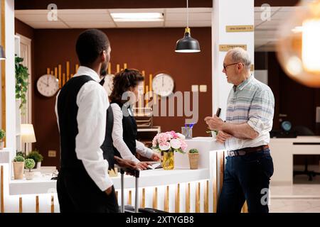 Un anziano anziano in pensione alla reception che effettua il check-in nella hall dell'hotel di prima qualità. Mentre il fattorino aiuta con i bagagli nell'area lounge, la portineria femminile assiste il viaggiatore anziano nella prenotazione della camera. Foto Stock