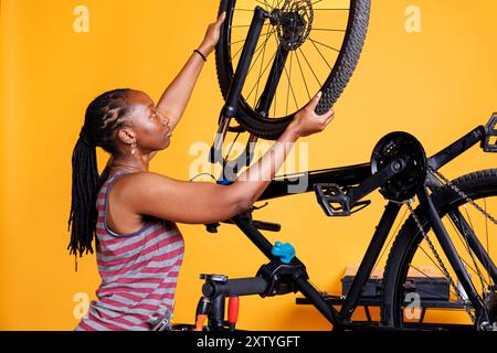 Meticolosa signora afroamericana ripara la moto rotta, regolando gli ingranaggi e fissando la ruota con precisione esperta. Donna nera sportiva che esamina e cambia pneumatico della bicicletta. Foto Stock