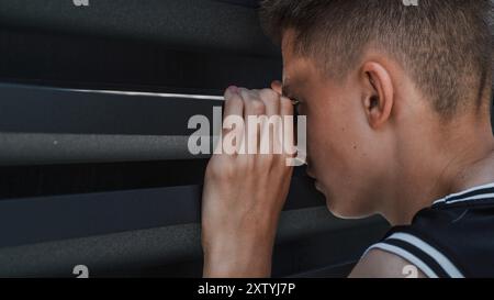 Movimento lento di un serio e concentrato adolescente caucasico che sbircia e guarda attentamente attraverso lo spazio tra la recinzione di ferro mentre si trova nel cortile della casa. Concetto di stile di vita. Primo piano. Foto Stock