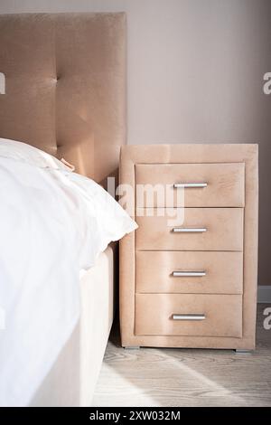 Comò beige con quattro cassetti vicino al letto. Camera ben organizzata con luce naturale. Foto Stock