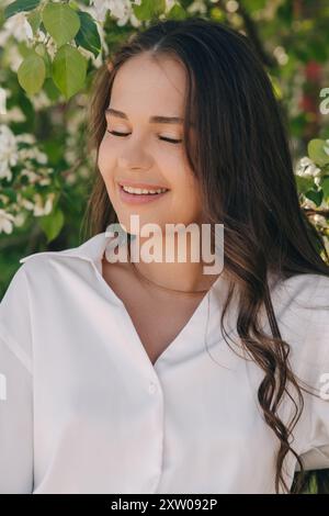 Ritratto di una bella ragazza con i capelli lunghi vicino a un albero in fiore in abiti bianchi alla moda Foto Stock