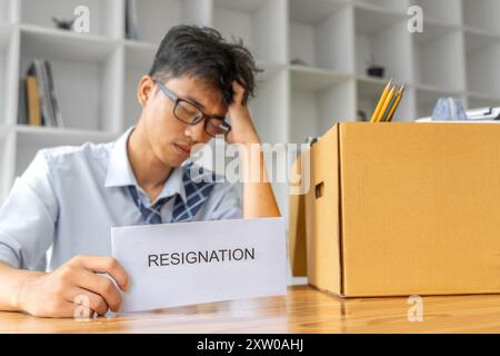 Triste giovane uomo d'affari seduto nel suo ufficio tenendo la sua lettera di dimissioni con una scatola piena di cose del suo ufficio accanto a lui Foto Stock