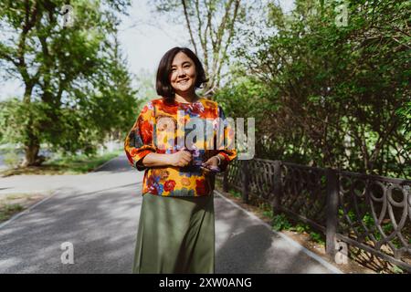 Un'elegante donna asiatica con un motivo floreale sorride sulla ringhiera del parco. Gioiosa giovane donna asiatica che si gode una passeggiata in un'estate Foto Stock