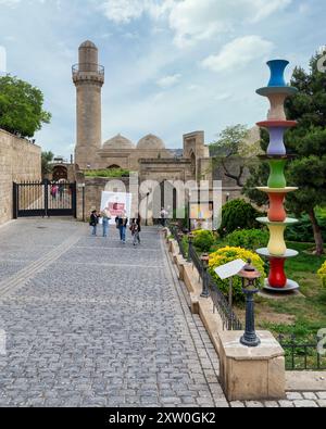 Baku, Azerbaigian - 5 maggio 2024: I turisti passeggiano attraverso il sentiero acciottolato che conduce alla storica Moschea dello Scià e al Palazzo Shirvanshah, con sculture colorate in primo piano Foto Stock