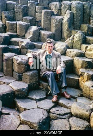 Uomo seduto su colonne di basalto e rocce a Giant's Causeway, Contea di Antrim, Irlanda del Nord, Regno Unito, 1960 circa Foto Stock