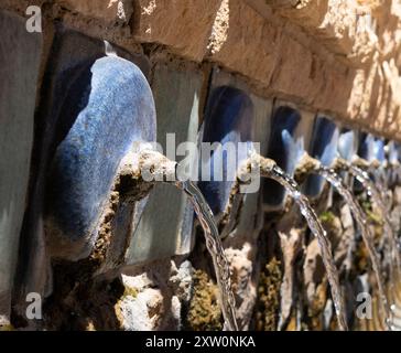 Vista dettagliata di diversi beccucci d'acqua su una fontana a parete in pietra, evidenziando le piastrelle blu e l'acqua che scorre. Foto Stock