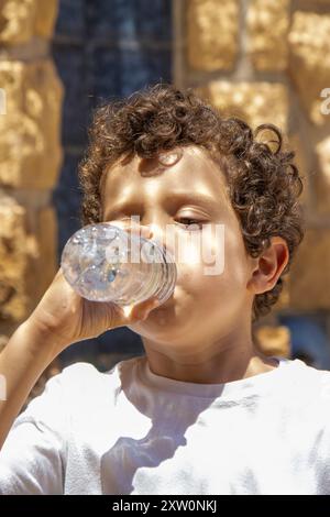 Un ragazzo beve l'acqua da una bottiglia mentre è in piedi all'aperto, mentre la luce del sole mette in risalto i suoi capelli ricci e il suo viso. Foto Stock