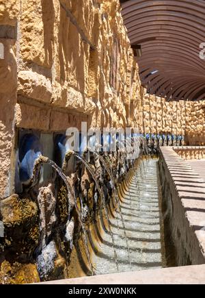 Vista dettagliata di una fontana a muro in pietra con diversi corsi d'acqua che scorrono sotto un pergolato di legno in una giornata di sole. Foto Stock