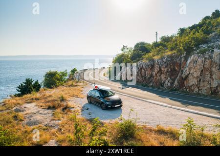 Auto nera con una tavola da paddleboard saldamente fissata in cima al tetto, parcheggiata sul lato dell'autostrada costiera con il mare scintillante Foto Stock