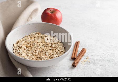 Avena cruda arrotolata in una ciotola di ceramica con bastoncini di cannella e mele su fondo testurizzato grigio chiaro Foto Stock