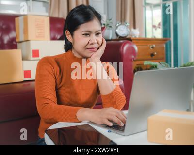 Donne asiatiche che lavorano in azienda SME online a casa il suo ordine di controllo dal computer portatile per il cliente e la consegna online per l'imballaggio pronto, controllando gli ordini da Foto Stock