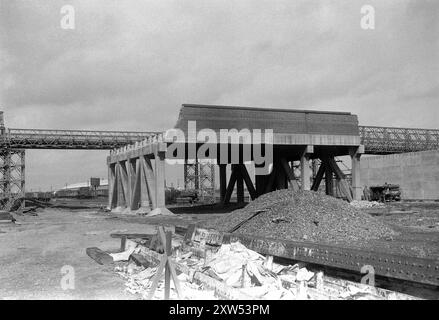 Inghilterra intorno al 1957 – Un ponte Bailey prefabbricato eretto sui binari ferroviari come sostituzione temporanea di un ponte stradale esistente, che è in fase di demolizione. Ci sono travi di ferro e un grande mucchio di macerie in primo piano. Gli operai utilizzano un compressore pneumatico e i carrelli ferroviari vengono immagazzinati in binari a distanza. Foto Stock